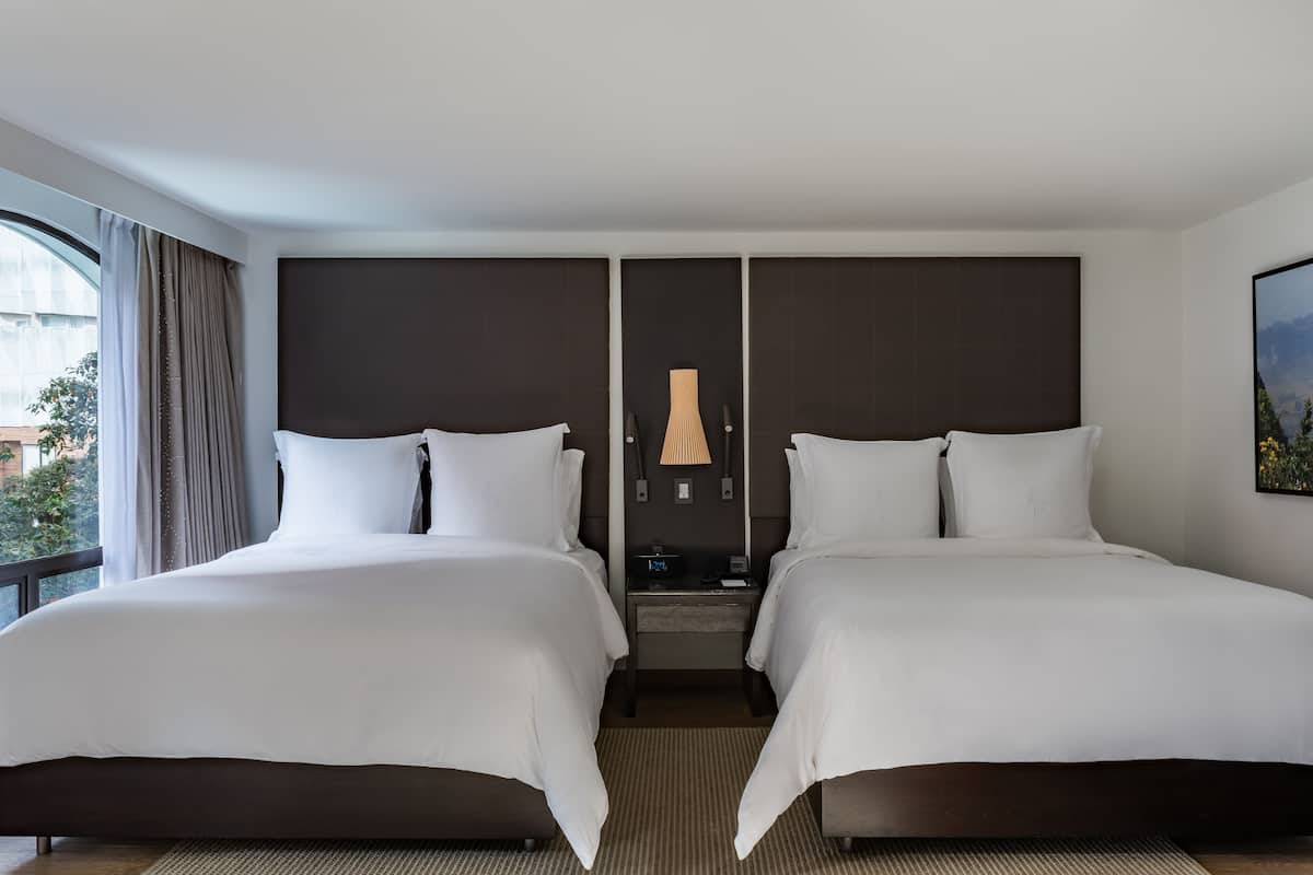 A hotel room with two beds, a nightstand, a lamp, and a window with curtains.