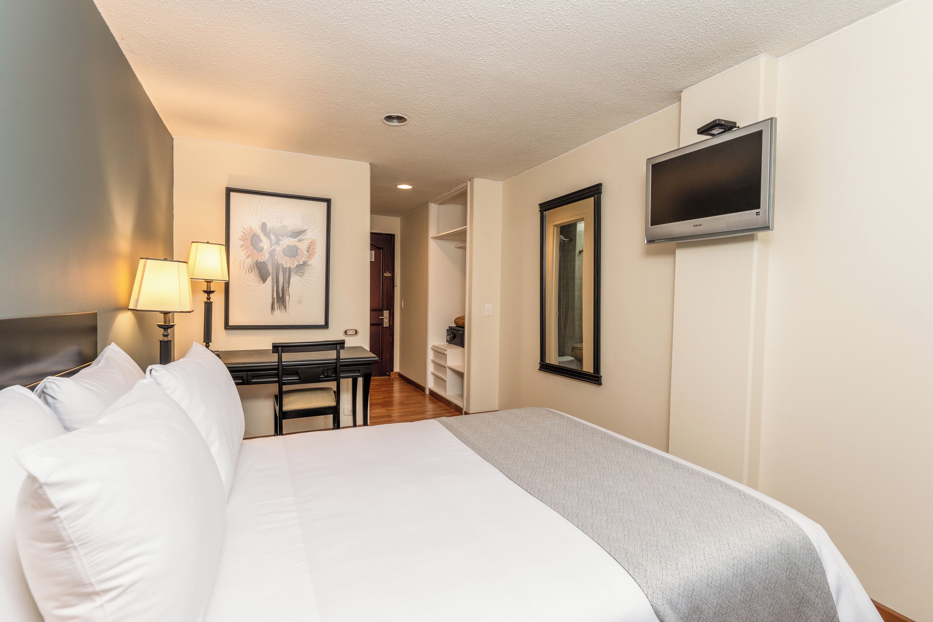 Een hotelkamer met een bed, bureau, stoel, lampen, een kunstwerk in een lijst en een televisie.