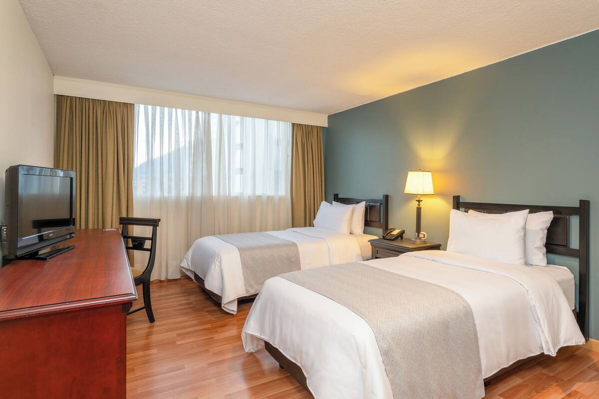 Habitación de hotel con dos camas, un escritorio con televisor, una lámpara y una ventana con cortinas.