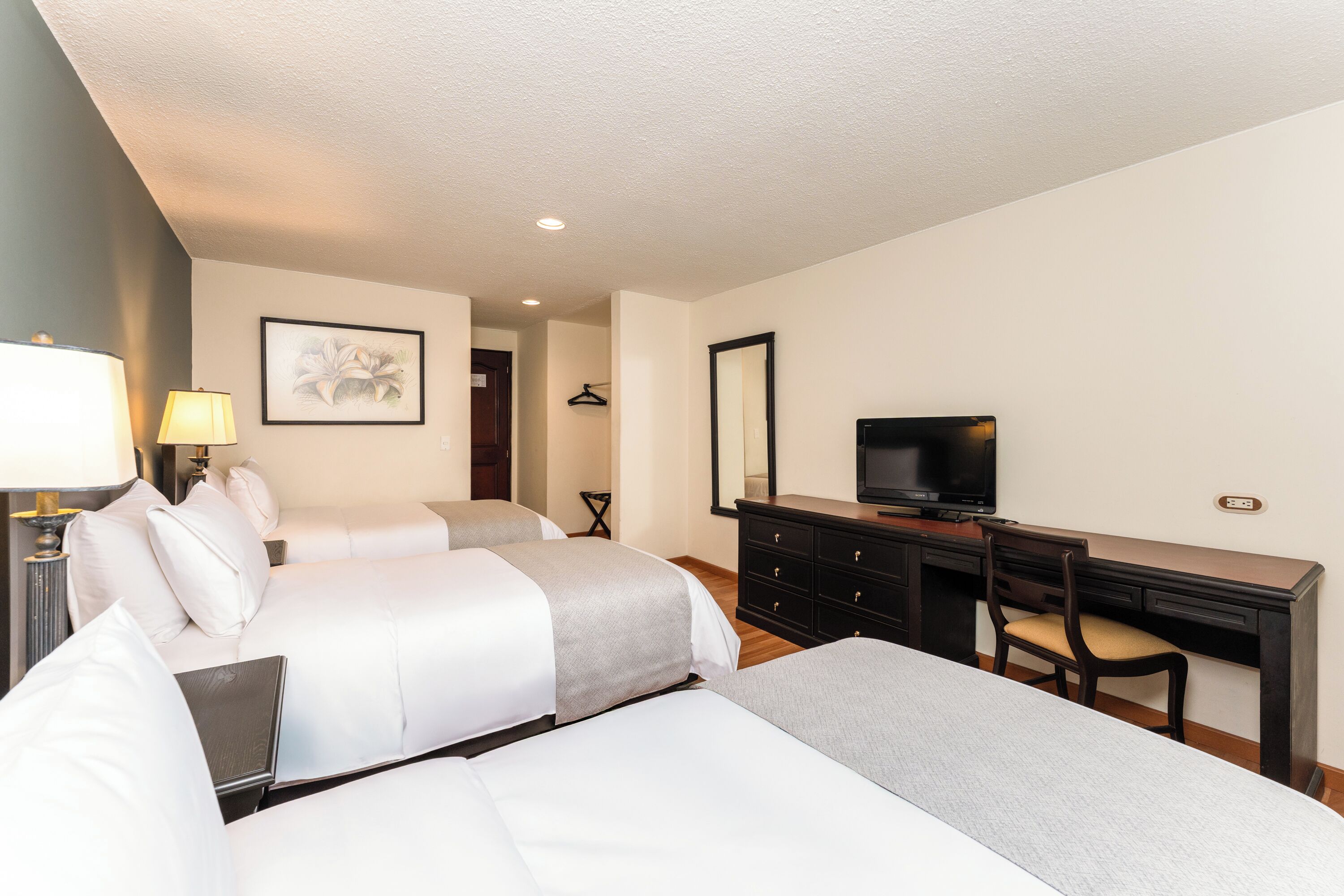 A hotel room with two beds, a desk, a chair, a TV, and a lamp.