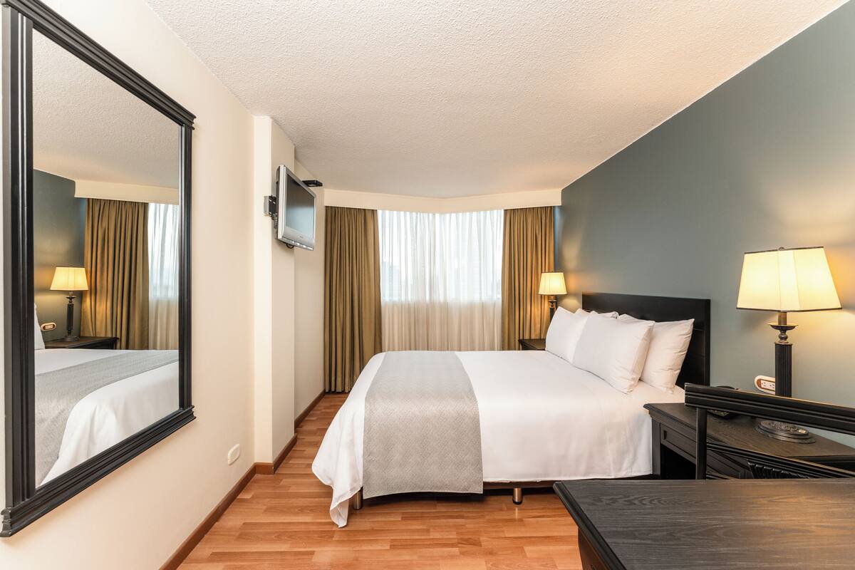 Una habitación de hotel con una cama, mesitas de noche, un televisor y un espejo grande.