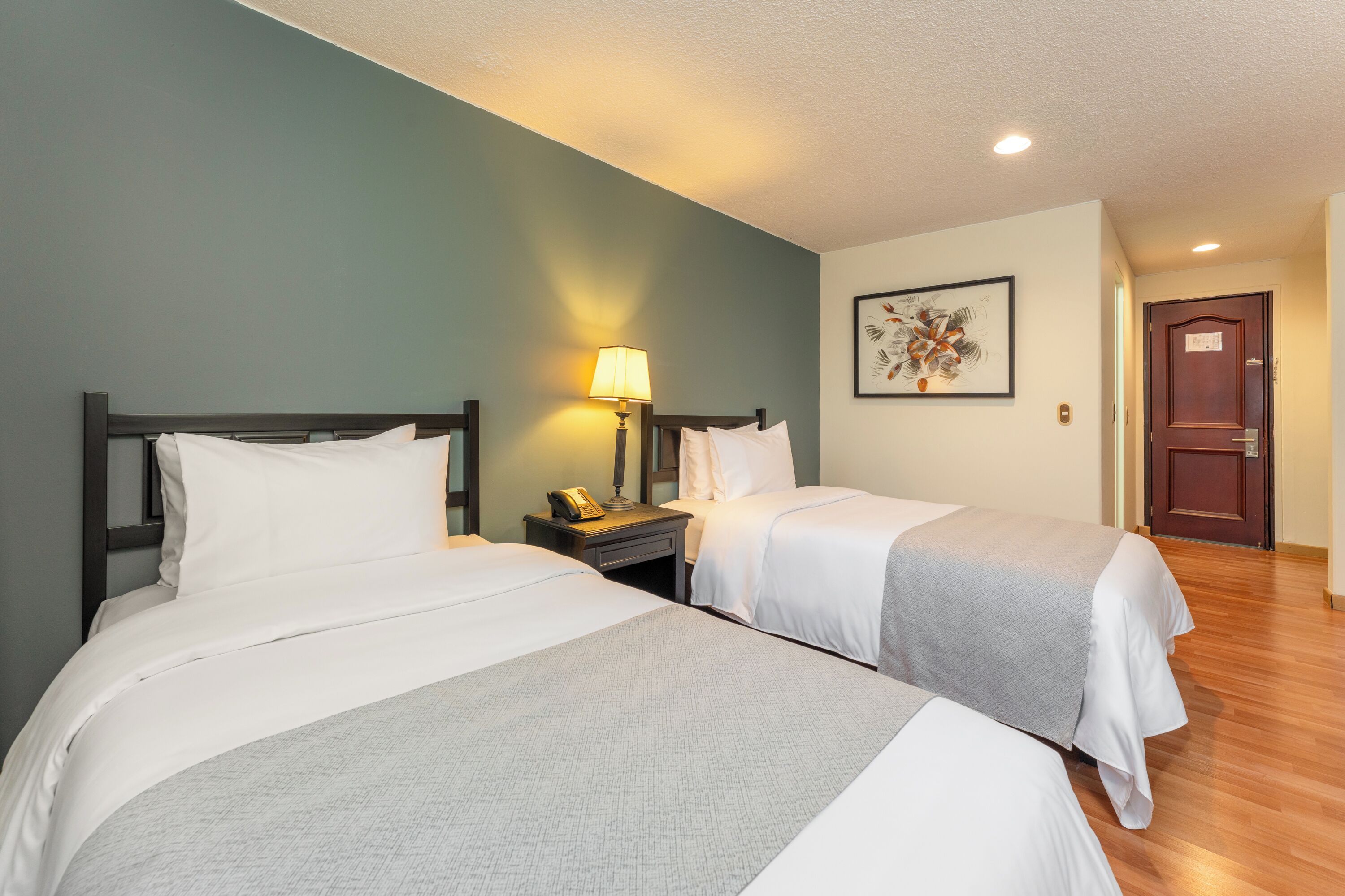 Ein Hotelzimmer mit zwei Betten, einem Nachttisch mit Lampe, einem gerahmten Bild an der Wand und einer Holztür.
