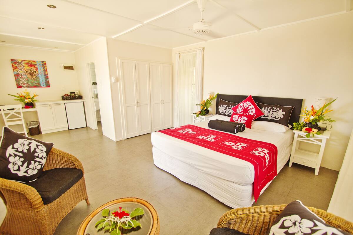 A hotel room with a bed, two nightstands, a wicker chair, a small table with a plant, and a kitchen area in the background.