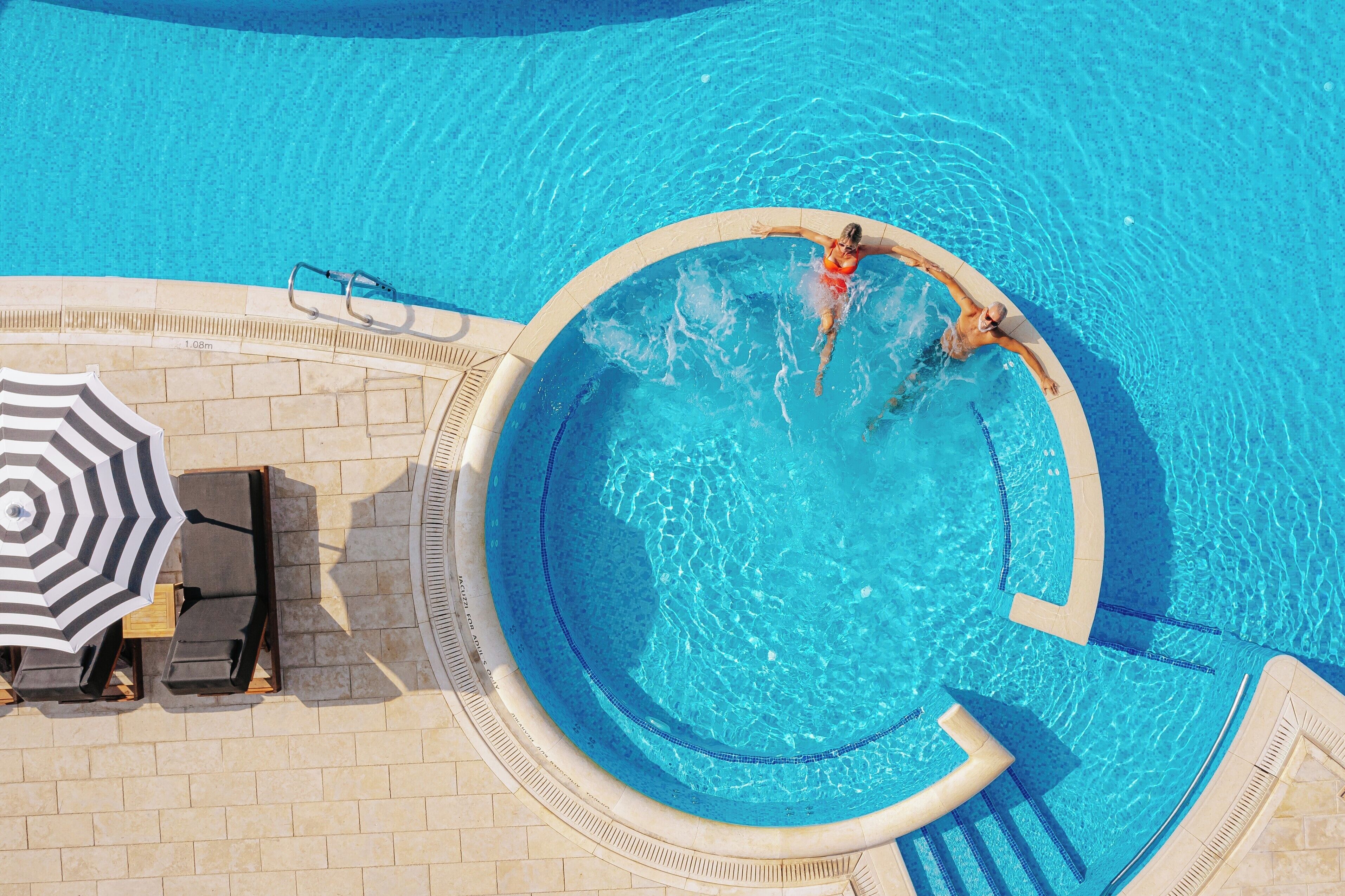 An aerial view of a swimming pool with two people in the water, a lounge area with chairs and an umbrella, and a tiled deck surrounding the pool.