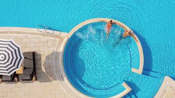 An aerial view of a swimming pool with two people in the water, a lounge area with chairs and an umbrella, and a tiled deck surrounding the pool.