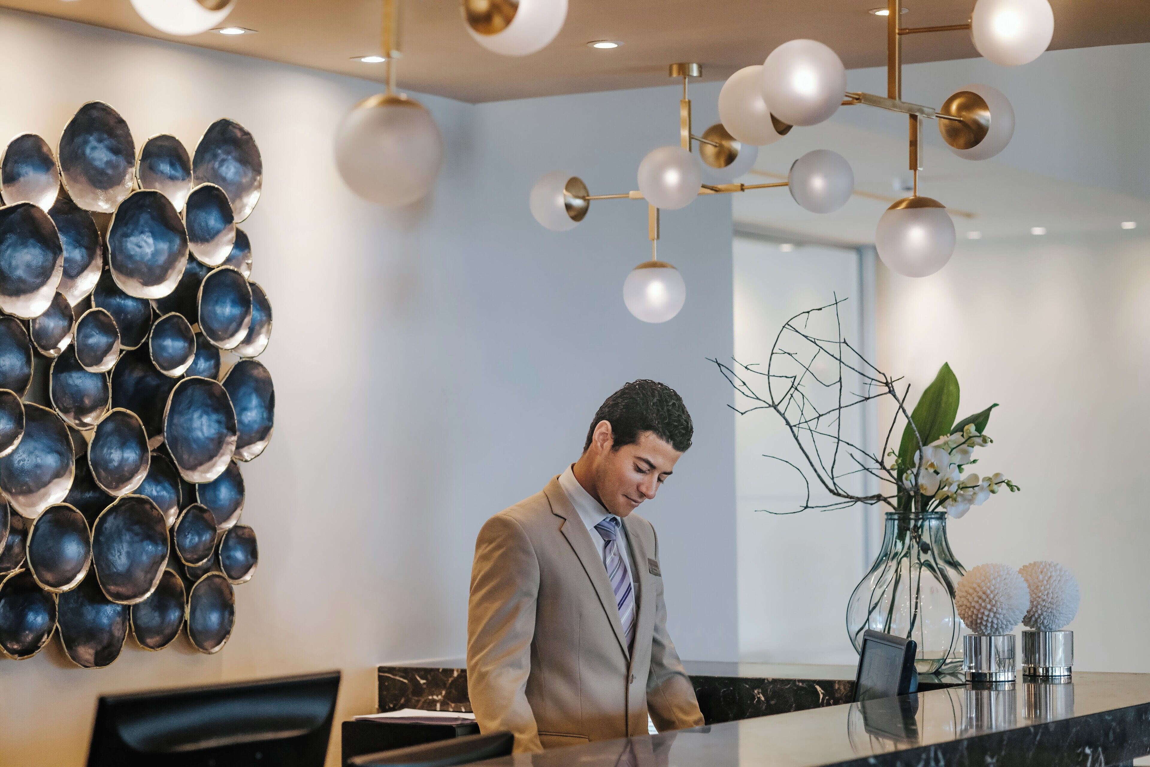 A modern reception desk with a glass top, a wall-mounted decorative metal piece, and a hanging light fixture.