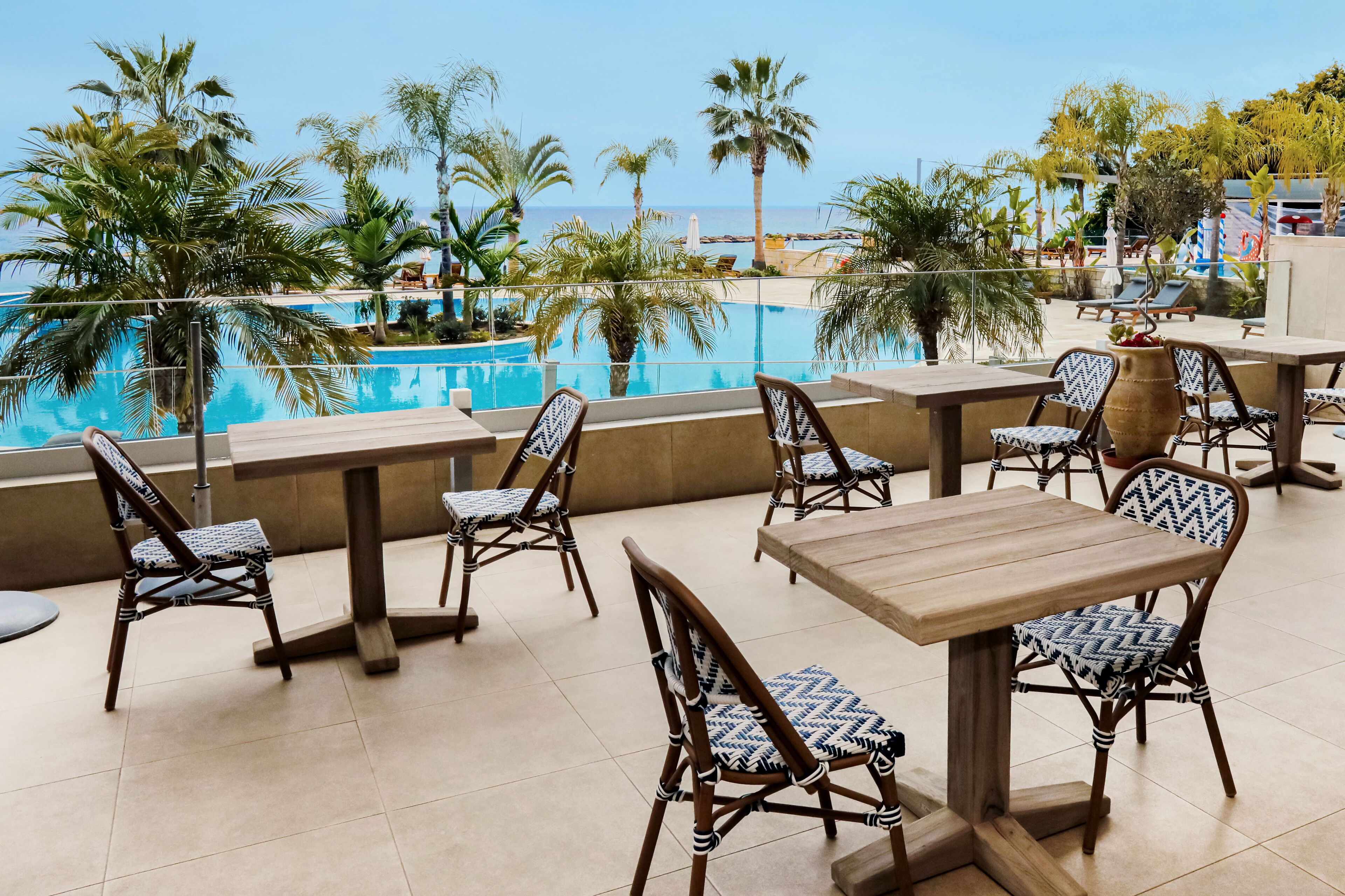 An outdoor dining area with a pool view, wooden tables and chairs, and palm trees.