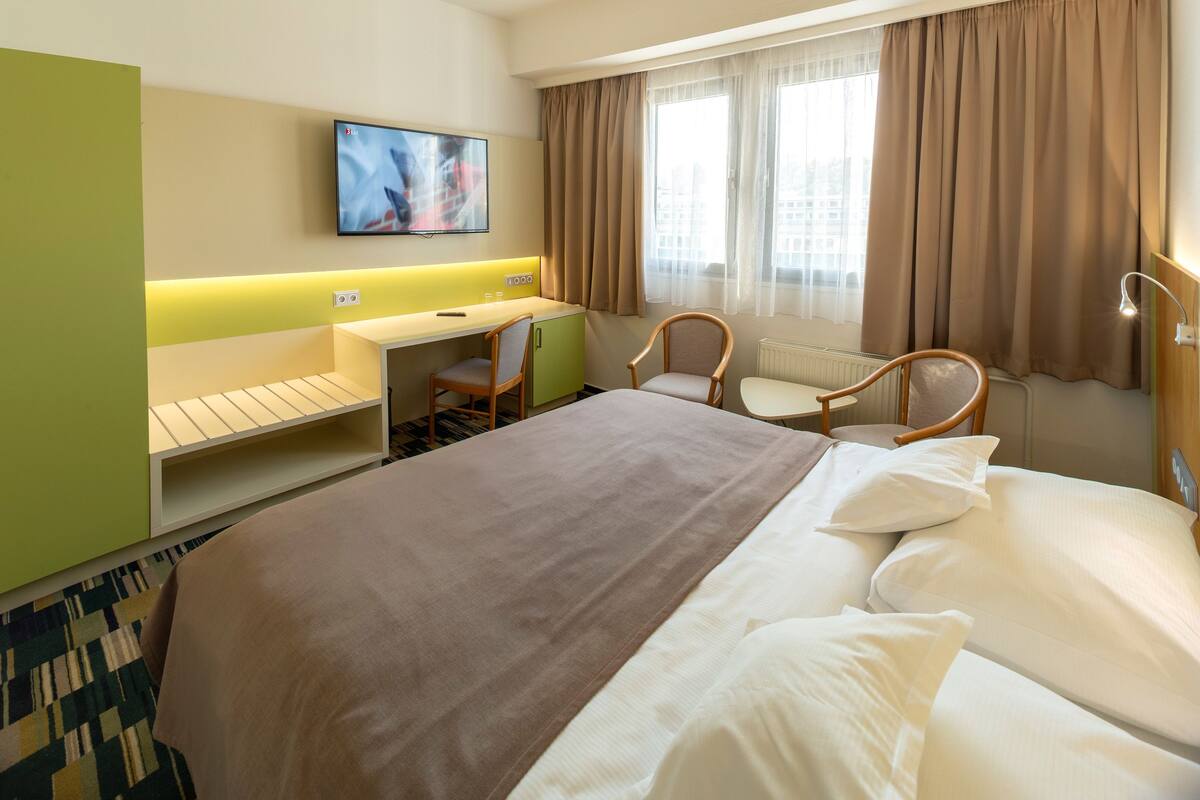 Una habitación de hotel con cama, escritorio, silla, televisor y ventana con cortinas.