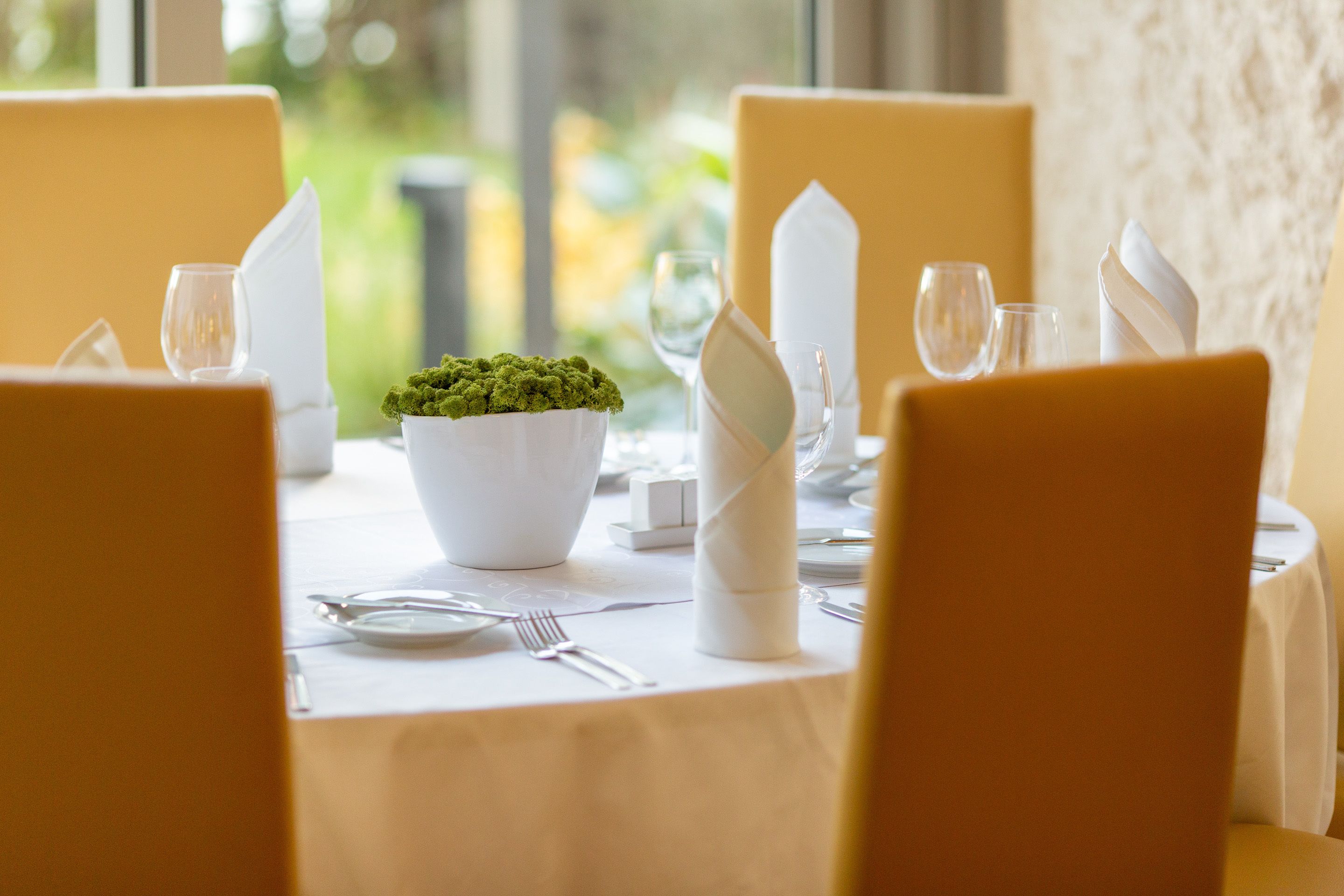 A dining table set with white tablecloth, yellow chairs, and a centerpiece of green foliage.