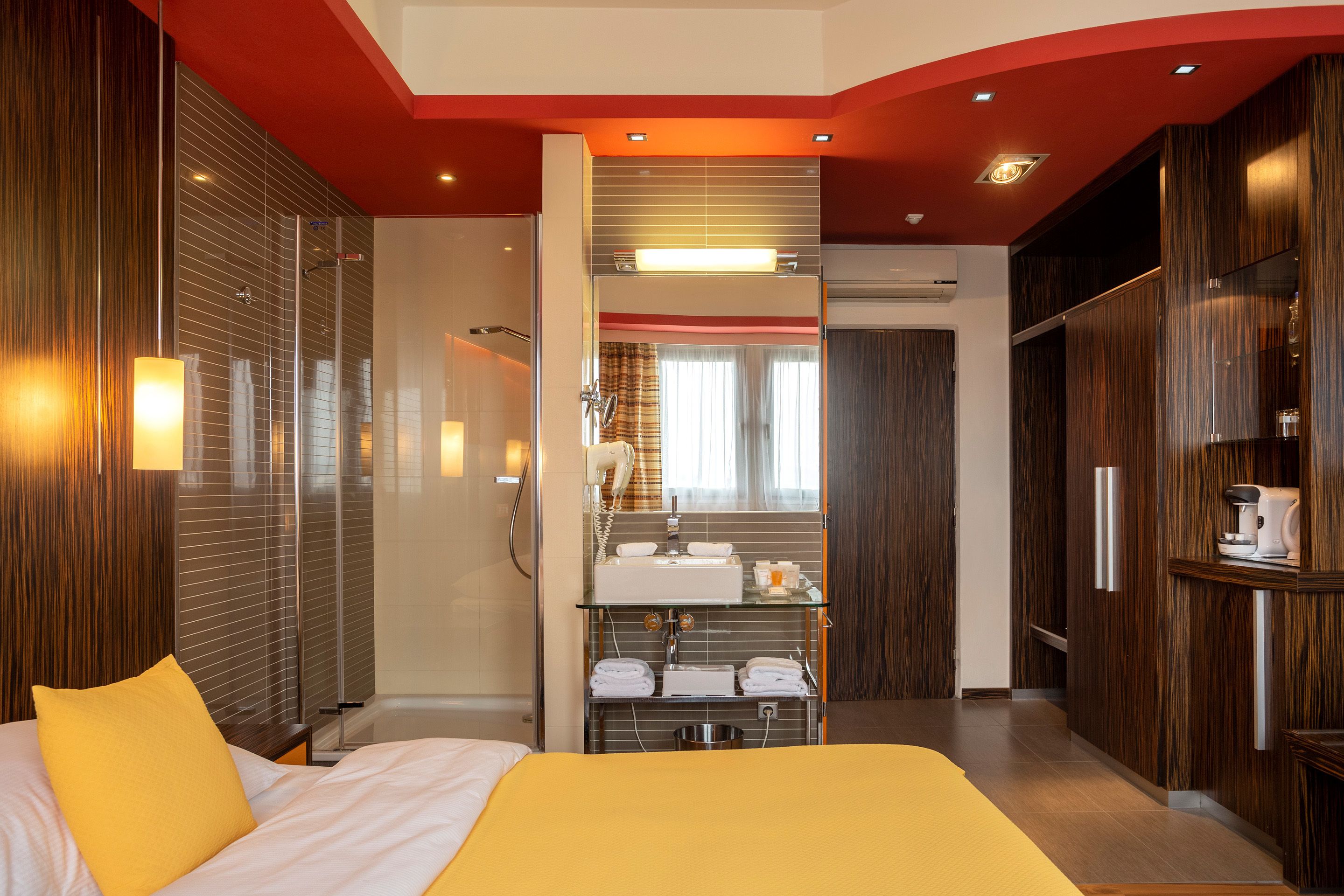 Una habitación de hotel moderna con una cama grande, cabina de ducha de vidrio, tocador con espejo y paneles de madera en las paredes.