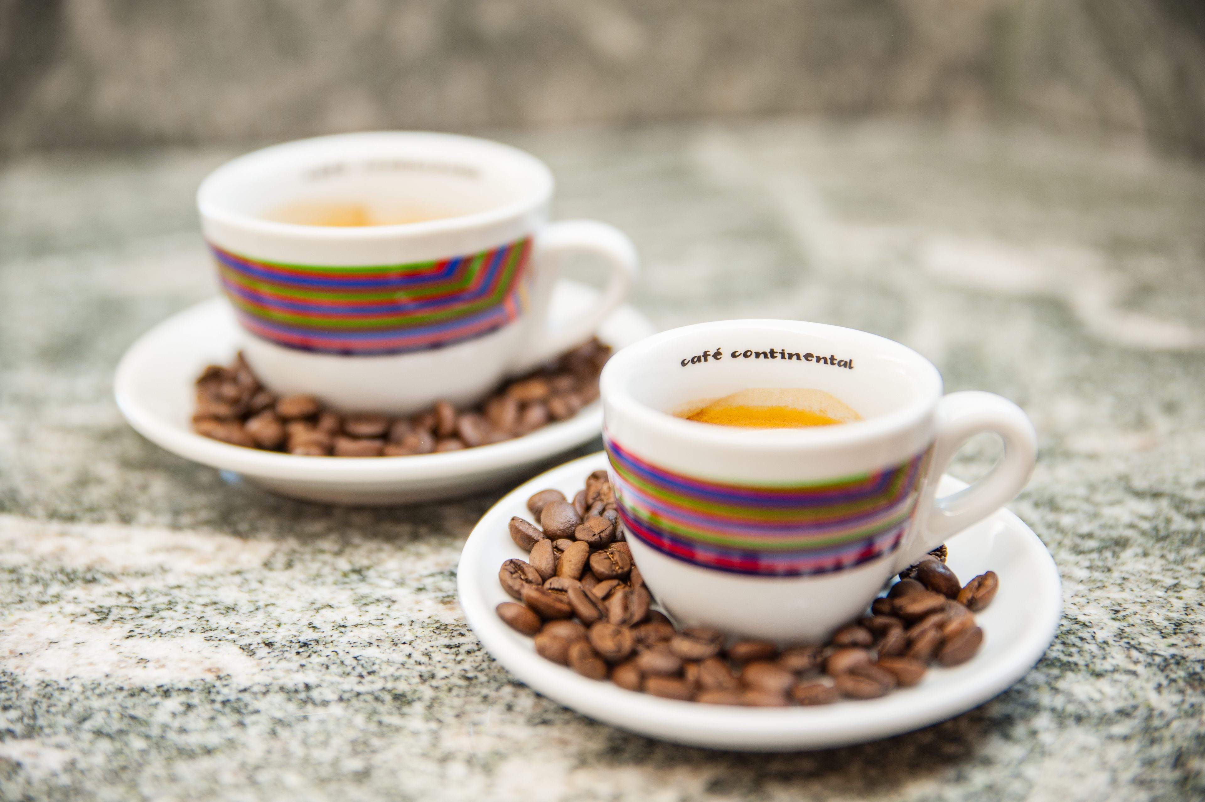 Two white cups with colorful stripes and the text "café continental" on them, placed on saucers with coffee beans around them.