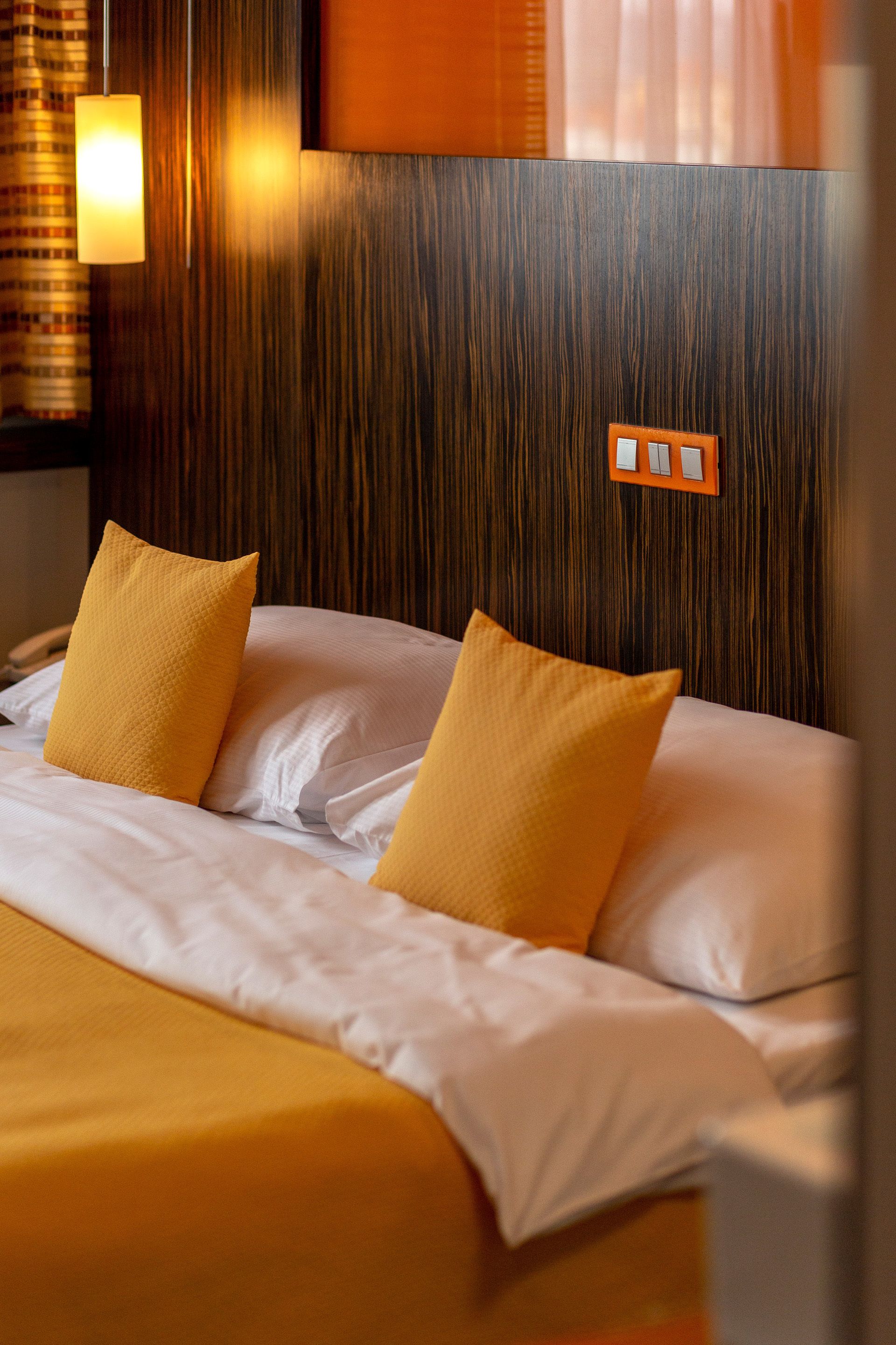 Habitación de hotel con cabecera de madera, dos almohadas amarillas, una colcha blanca y una lámpara fijada a la pared.