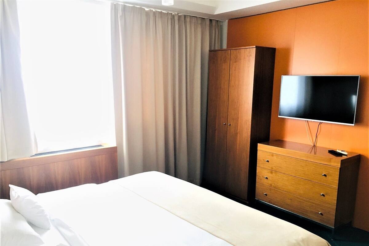 Una habitación de hotel con cama, armario, tocador, televisor y ventana con cortinas.