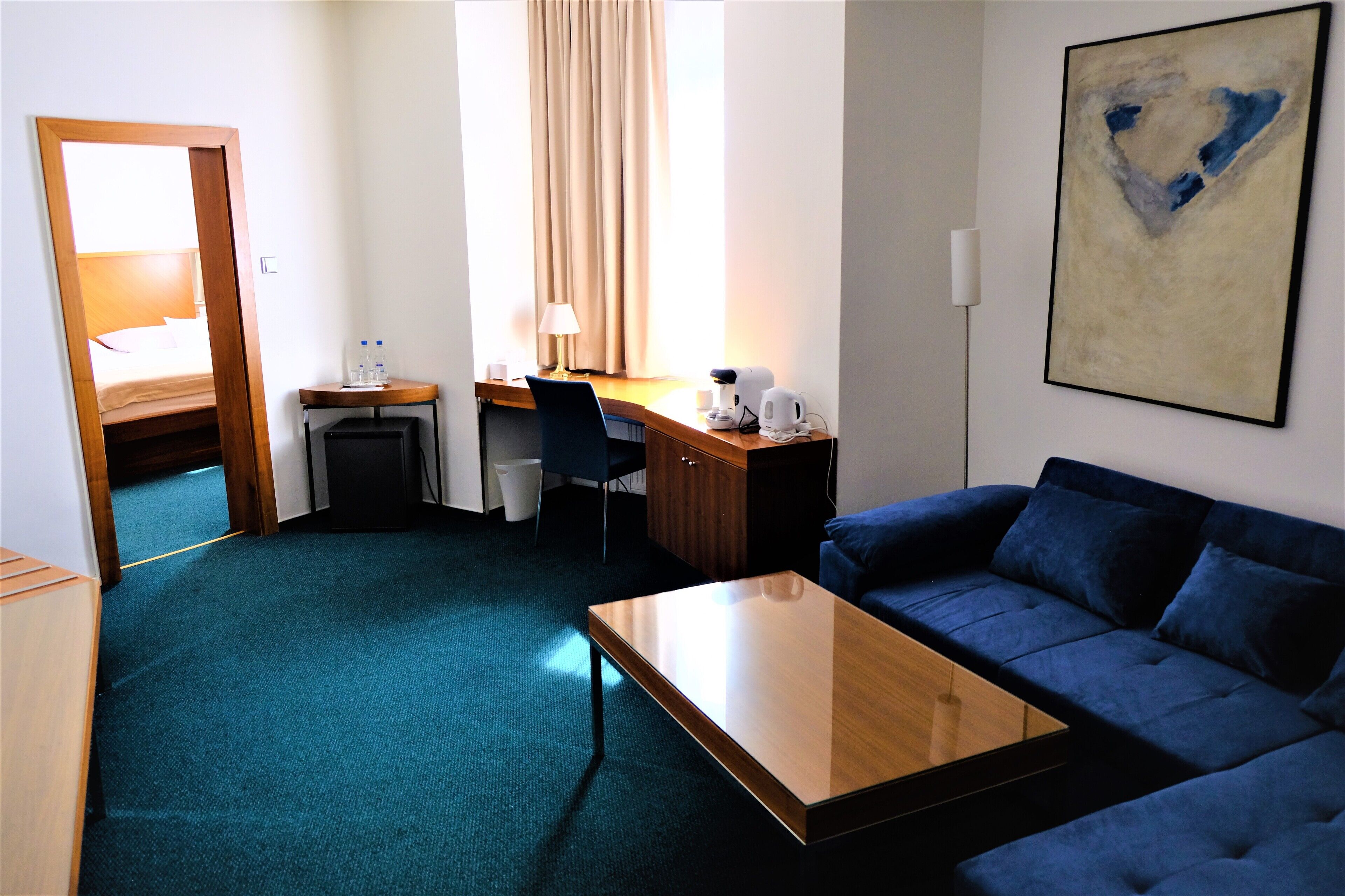 Habitación de hotel con sofá azul, una mesa de centro de madera, un escritorio con silla, una lámpara y un cuadro en la pared.