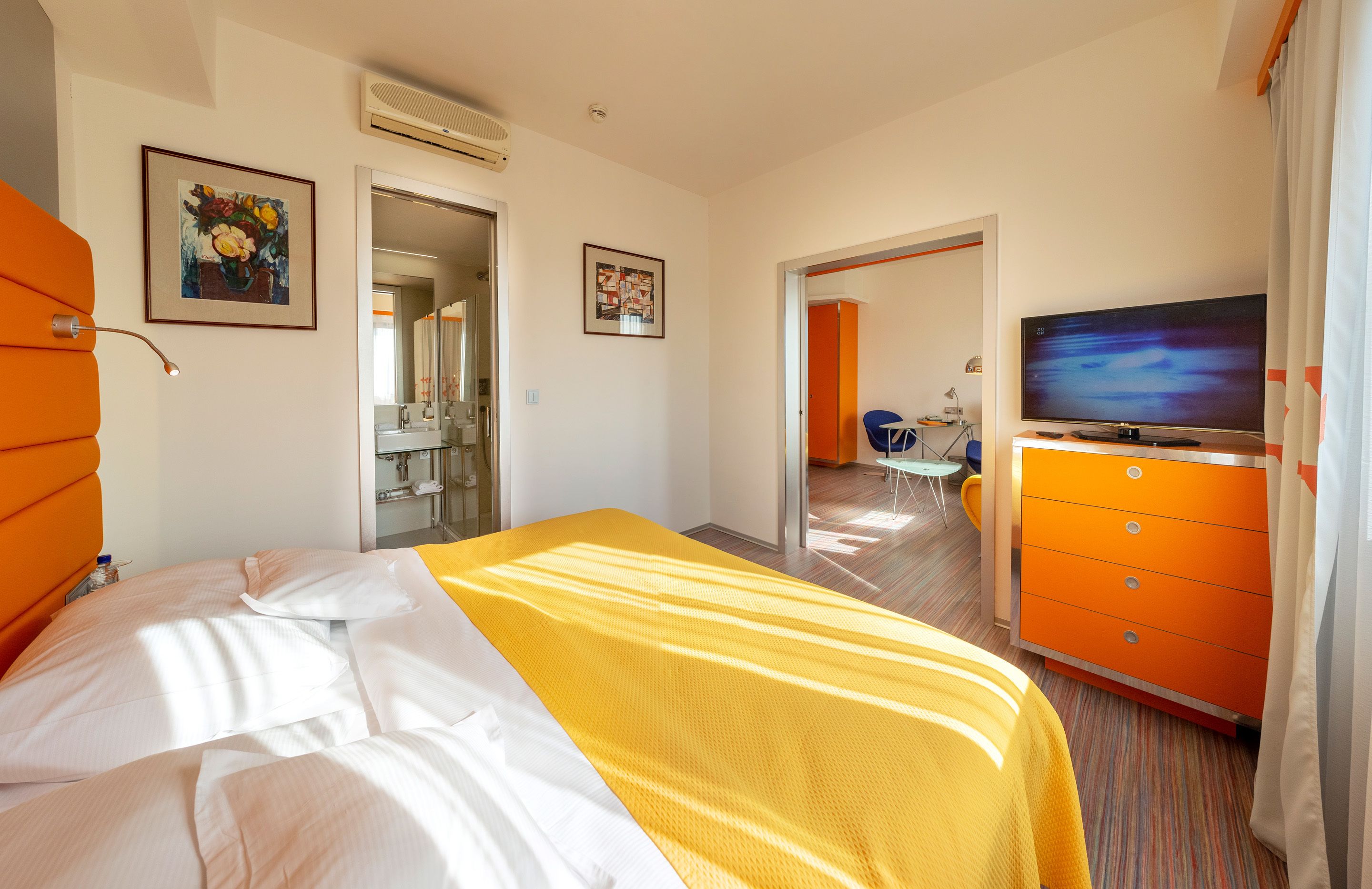 A hotel room with a bed, a TV, an orange dresser, and a bathroom visible through an open door.