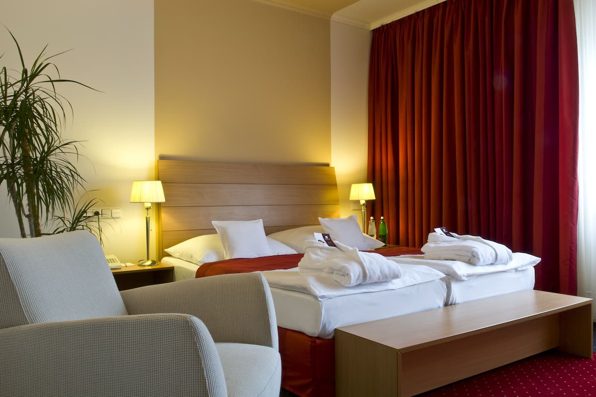 Habitación de hotel con cama, dos mesitas de noche, una silla, un escritorio, una lámpara, una planta y una ventana con cortinas rojas.