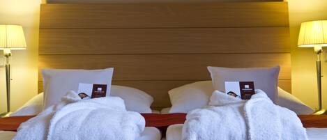 A hotel room with two beds, each with white linens, pillows, and a neatly folded towel. There are two bedside lamps and a wooden headboard.