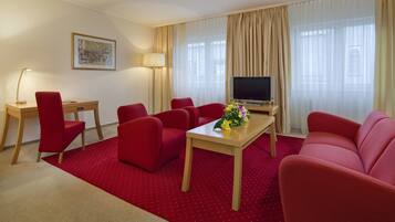 Quarto de hotel com carpete vermelho, escrivaninha de madeira, televisão, sofá e abajur.
