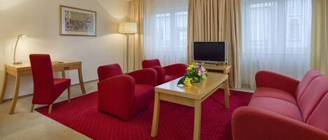 Habitación de hotel con alfombra roja, un escritorio de madera, televisión, sofá y lámpara.
