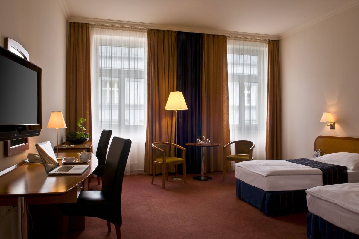 Habitación de hotel con escritorio, dos sillas, televisión, dos camas, lámpara y ventana con cortinas.