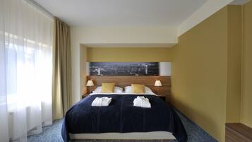 A hotel room with a large bed, two bedside lamps, a cityscape painting on the headboard, and a view of the city through sheer curtains.