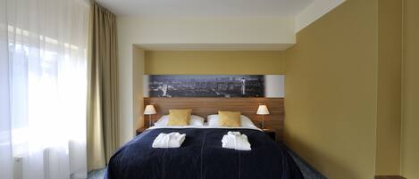 A hotel room with a large bed, two bedside lamps, a cityscape painting on the headboard, and a view of the city through sheer curtains.
