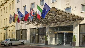 A hotel entrance with multiple flags, including the UK, Italy, and EU flags, and a car parked in front.