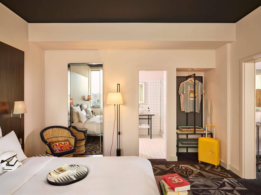 A hotel room with a bed, a chair, a lamp, a suitcase, and a bathroom visible through an open door.