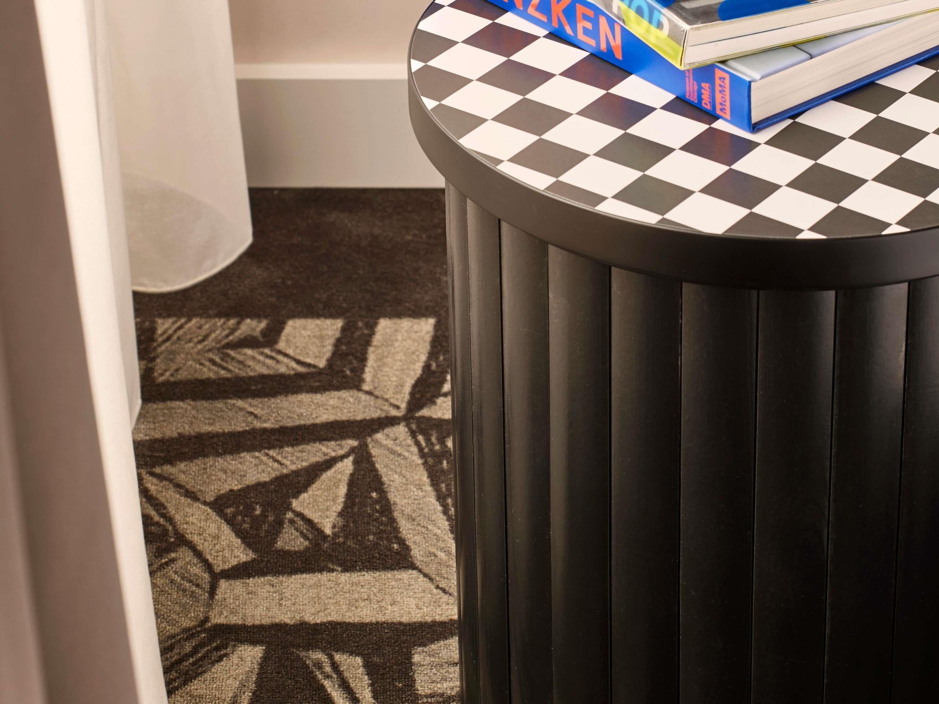 A round table with a checkered top, a black trash bin, and a patterned carpet.