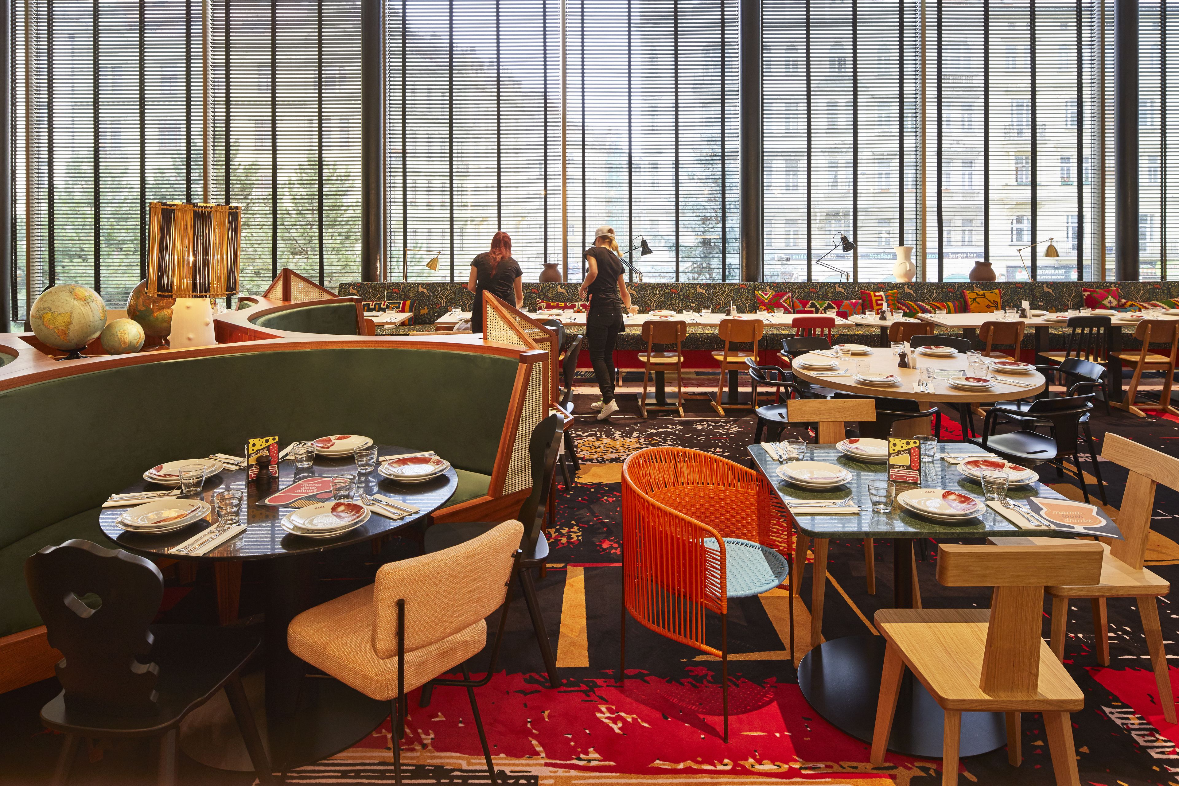 Een ruime eethoek met meerdere tafels en stoelen, grote ramen met zonwering en diverse gedekte tafels.