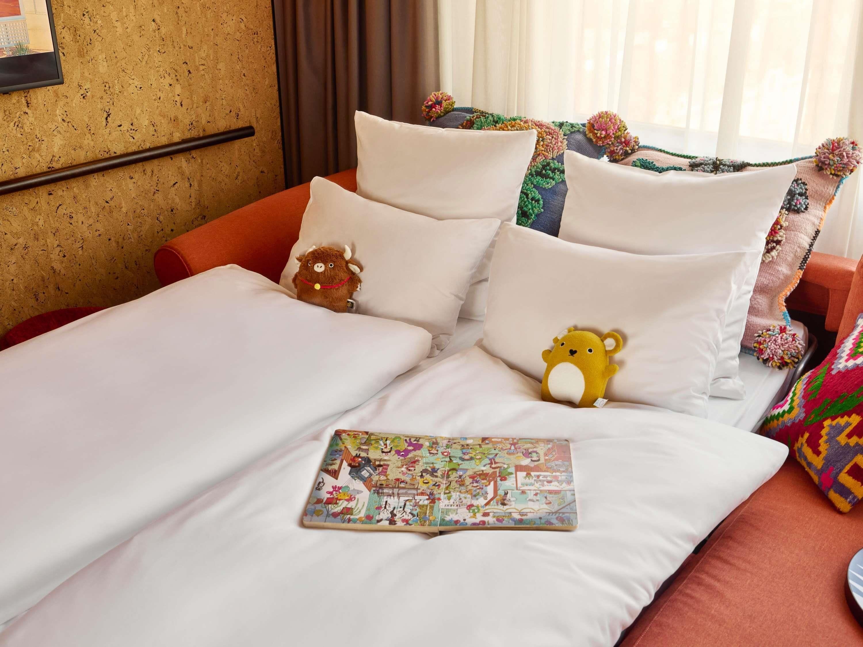 A bed with white linens, two plush toys, and a colorful book.