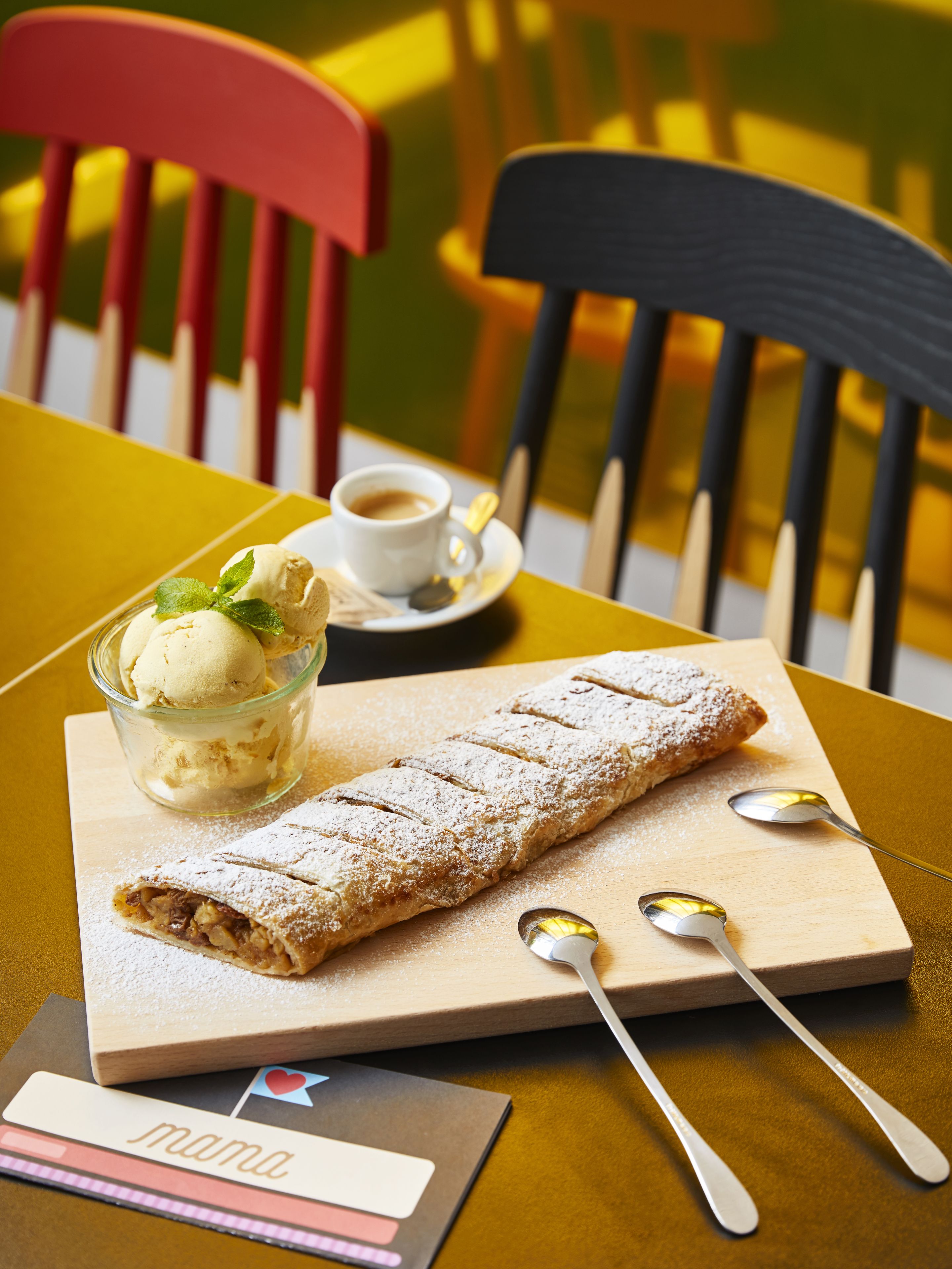 Un dessert composé d’une pâtisserie et d’une boule de glace, le tout présenté sur une planche en bois, avec des cuillères et une tasse de café sur une table.