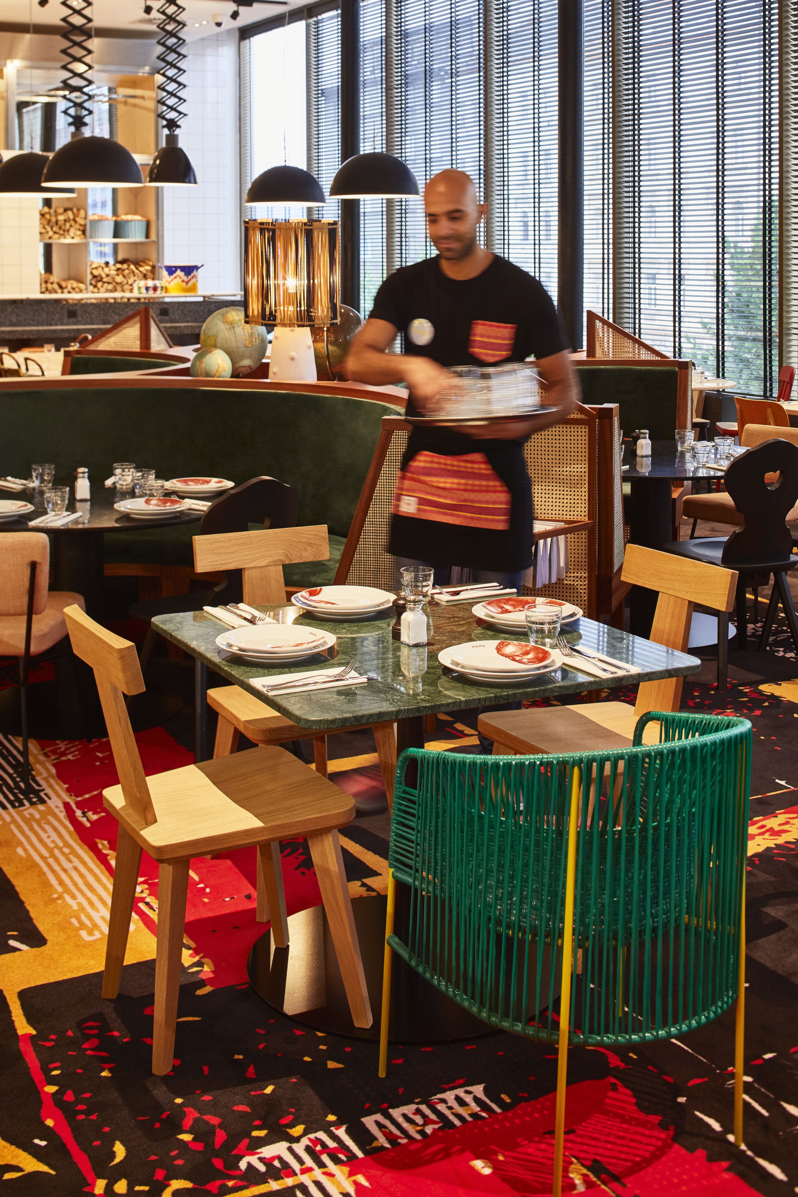 A restaurant with wooden chairs and tables, a waiter carrying a tray, and a large window in the background.
