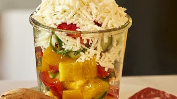Ein Glasbehälter mit einem Schichtsalat, der Gemüse und Käse enthält, zusammen mit Fladenbrot auf einem Teller.