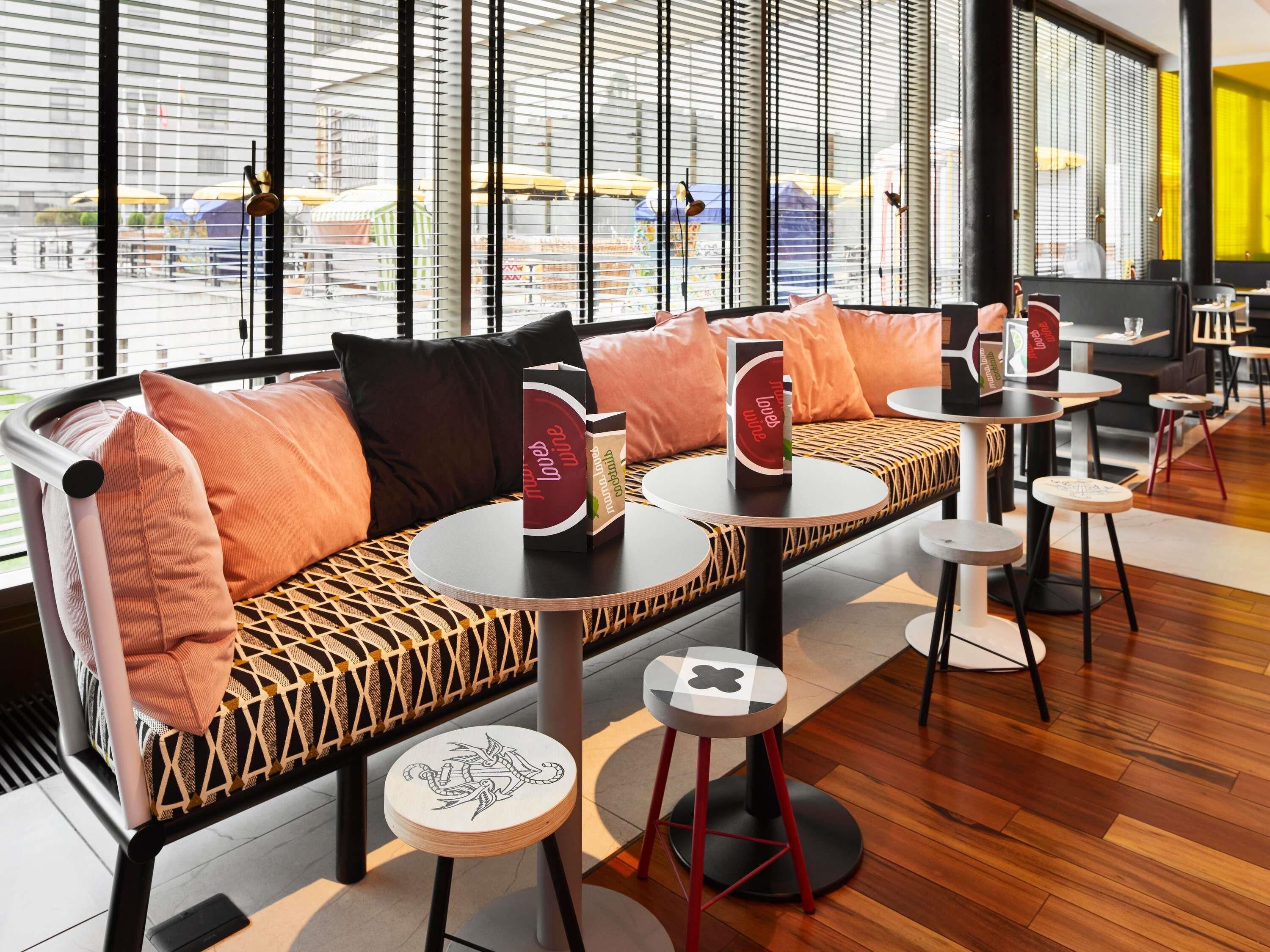 A modern seating area with a sofa, round tables, and stools, featuring a view through large windows.