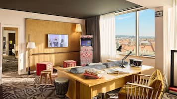 A hotel room with a large window, a television, a desk with books, a drumming stool, and a patterned carpet.