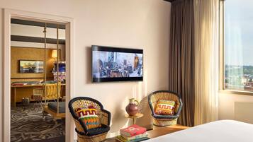 Una habitación de hotel con una cama, dos sillas de mimbre, un televisor en la pared y vistas de la ciudad a través de la ventana.