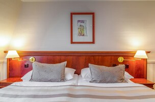 Quarto de hotel com cabeceira de madeira, duas luminárias de apoio, um quadro na parede e cama com roupa de cama listrada e almofadas estampadas.