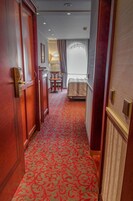 Un corridoio d'albergo con tappeto rosso a motivi decorativi, porte in legno, un letto, un tavolo e una finestra con tende.