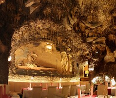 Restaurante com interior que lembra uma caverna, apresentando estalactites e uma grande pintura rupestre na parede. Mesas dispostas com velas e taças de vinho.