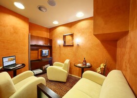 Quarto com um laptop sobre uma mesa, duas cadeiras amarelas, uma mesinha de centro com uma garrafa e um copo, e um espelho na parede.