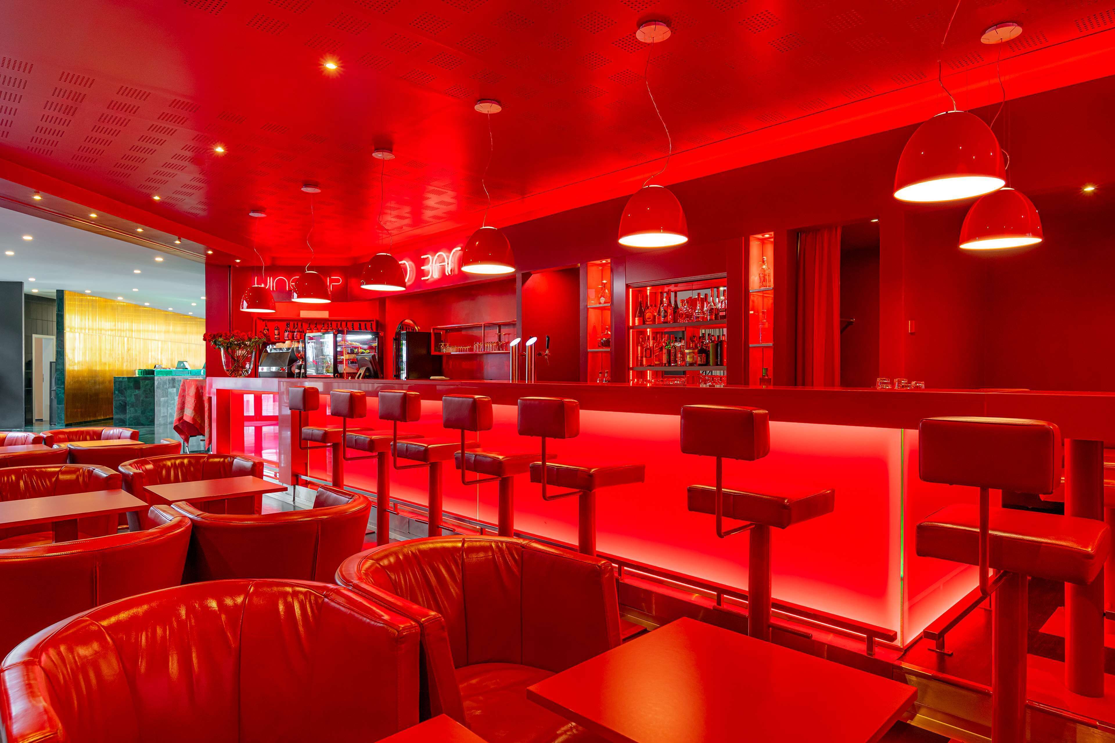 Interior de un restaurante con decoración roja, con cabinas y mesas de cuero, una zona de bar e iluminación colgante.