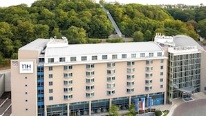 A multi-story hotel with a modern design, featuring a prominent "NH Hotels" sign, a covered entrance, and a scenic view of a forested hill. - NH Prague City (Prague)