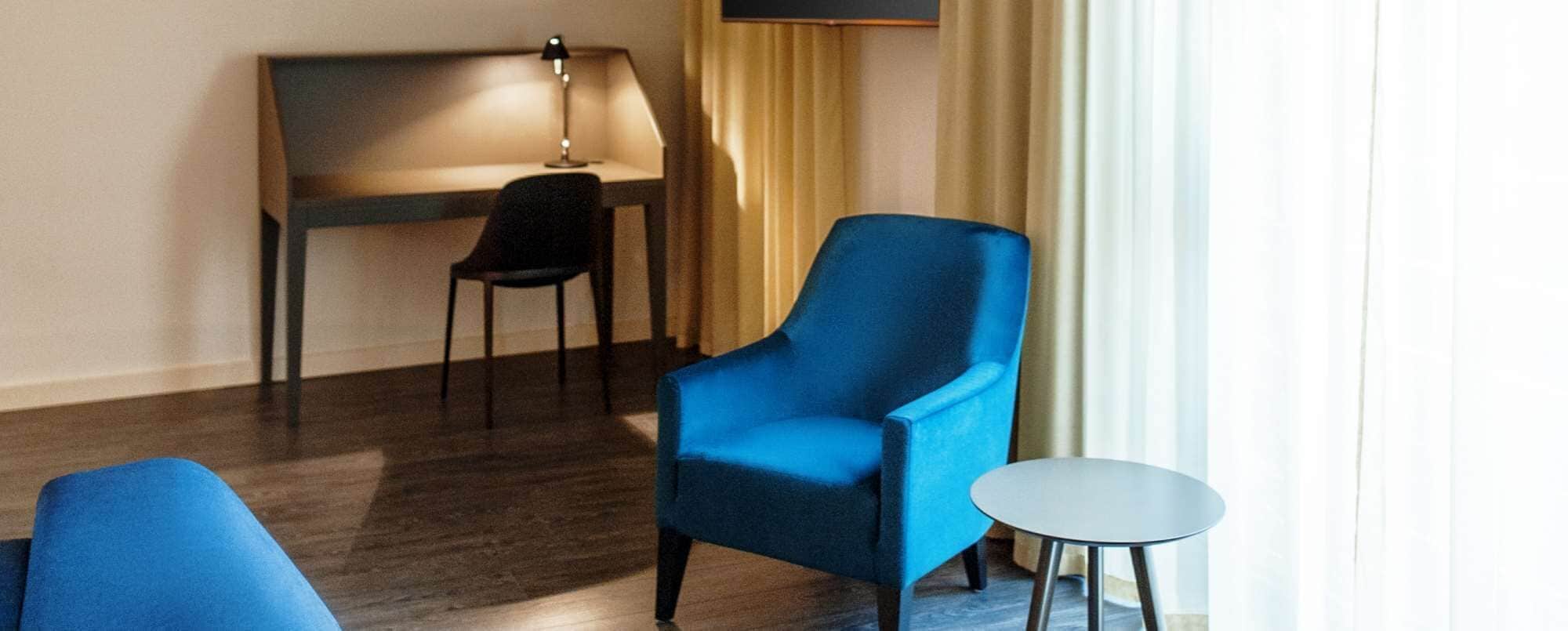 A hotel room with a blue armchair, a small round table, a desk with a lamp, and a television on the wall.