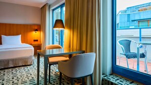 A hotel room with a bed, a table, two chairs, a lamp, and a view of the outside building through large windows. - NH Prague City (Prague)