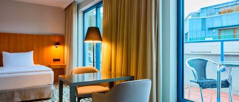 A hotel room with a bed, a table, two chairs, a lamp, and a view of the outside building through large windows.