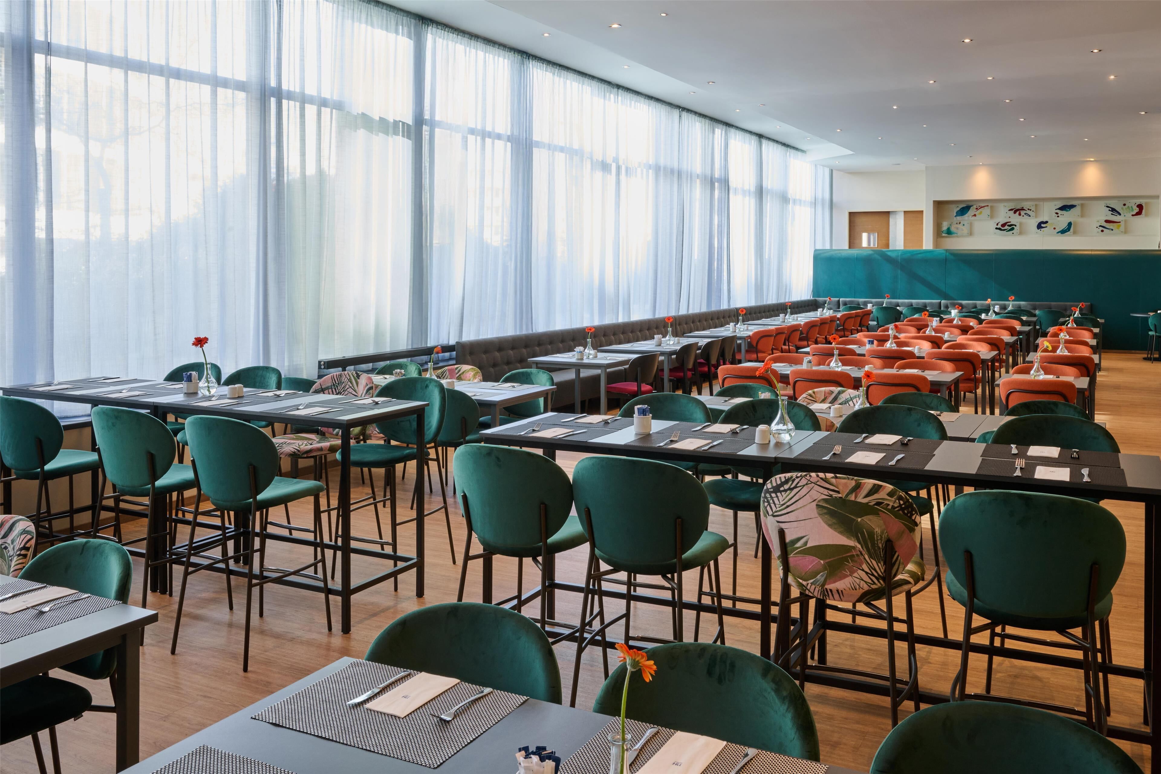 A spacious dining area with rows of tables and chairs, large windows with sheer curtains, and a teal-colored wall with artwork.
