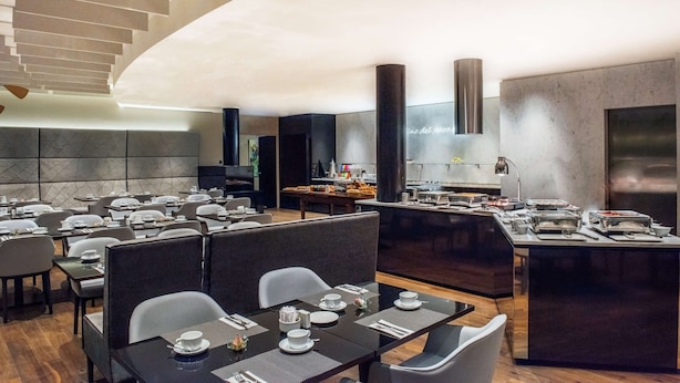 A modern restaurant with a long dining area, high ceilings, and a kitchen visible in the background. - NH Prague City (Prague)