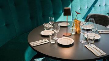 A round dining table set with plates, glasses, and cutlery, against a teal upholstered wall.