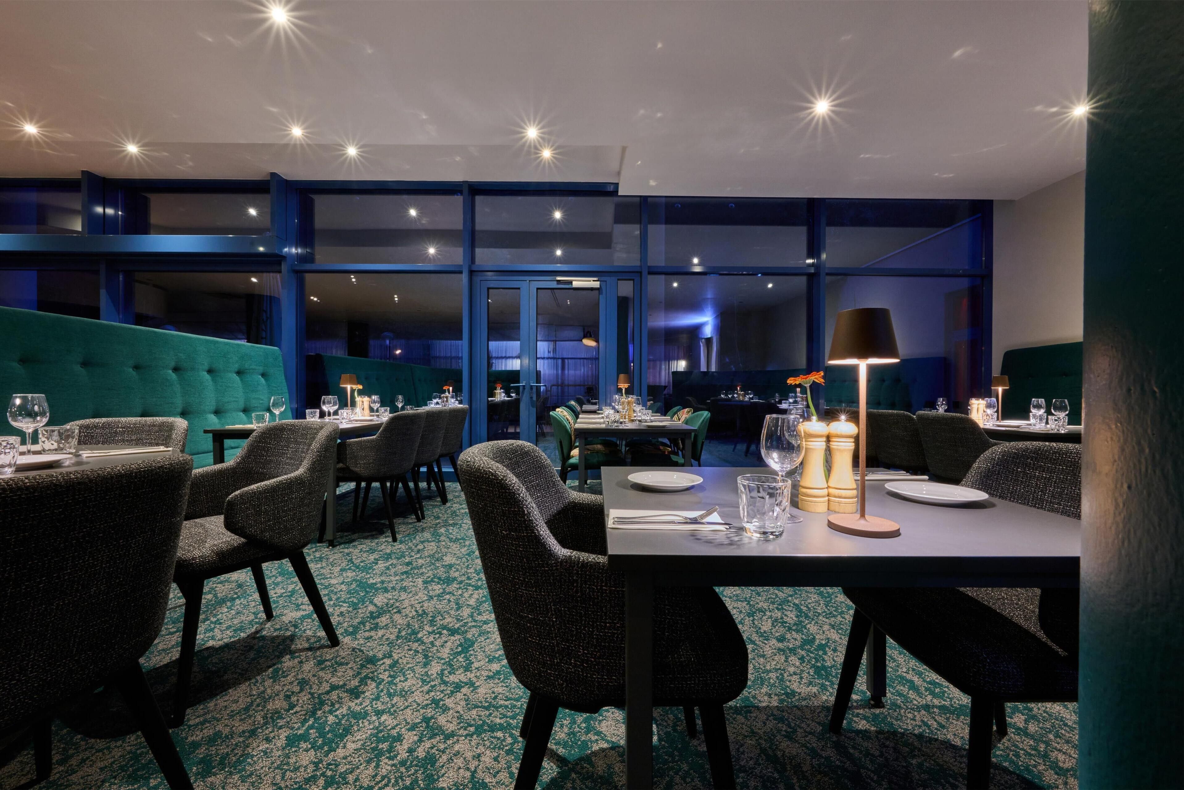 Un restaurante con un interior moderno, que cuenta con una banca tapizada en color aguamarina, sillas oscuras y mesas dispuestas con platos y vasos.